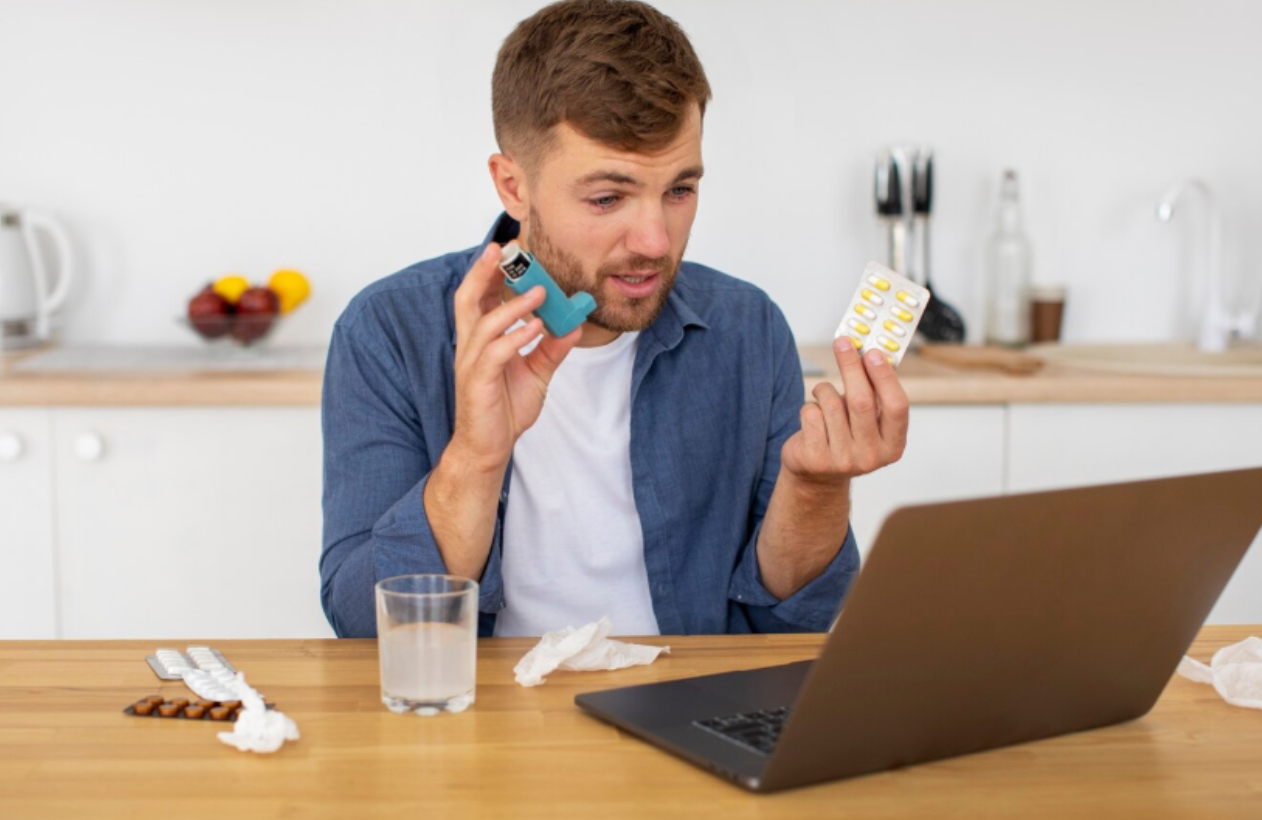 A man sitting at a kitchen table looking uncertain while reading a blister pack of pills and holding an inhaler, with his laptop open — representing caution when buying supplements online