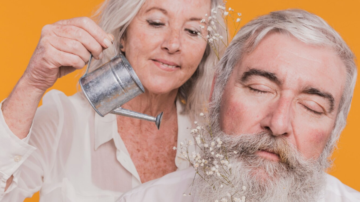 An older woman playfully pretends to water an older man’s beard with a small watering can, both smiling, symbolizing natural aging, hydration, and wellness.