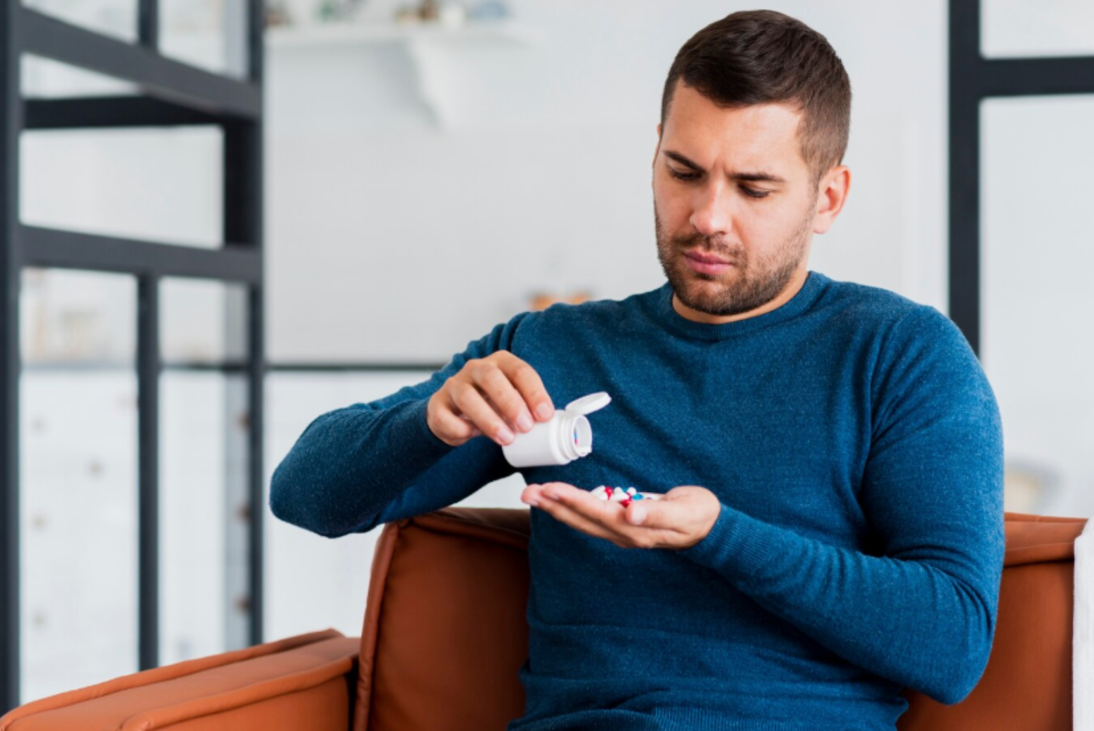 Man in his 30s examining a handful of colorful supplements while seated indoors, appearing uncertain about his daily routine.