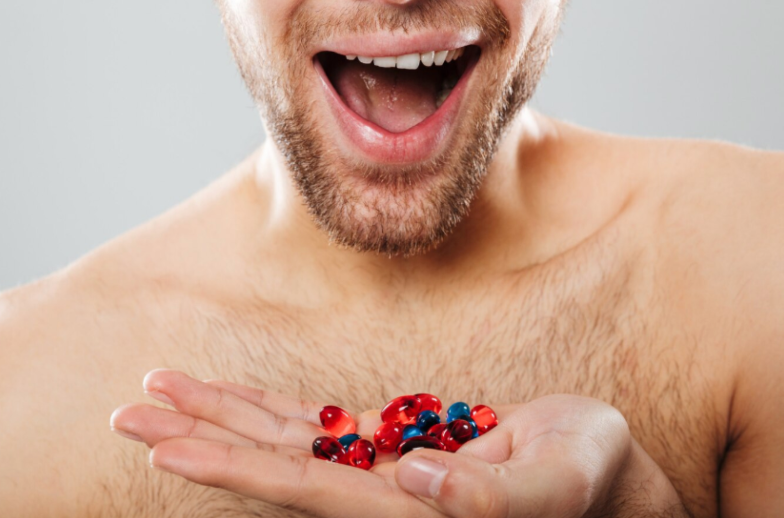 Close-up of a shirtless man smiling with a handful of red and blue supplement capsules.