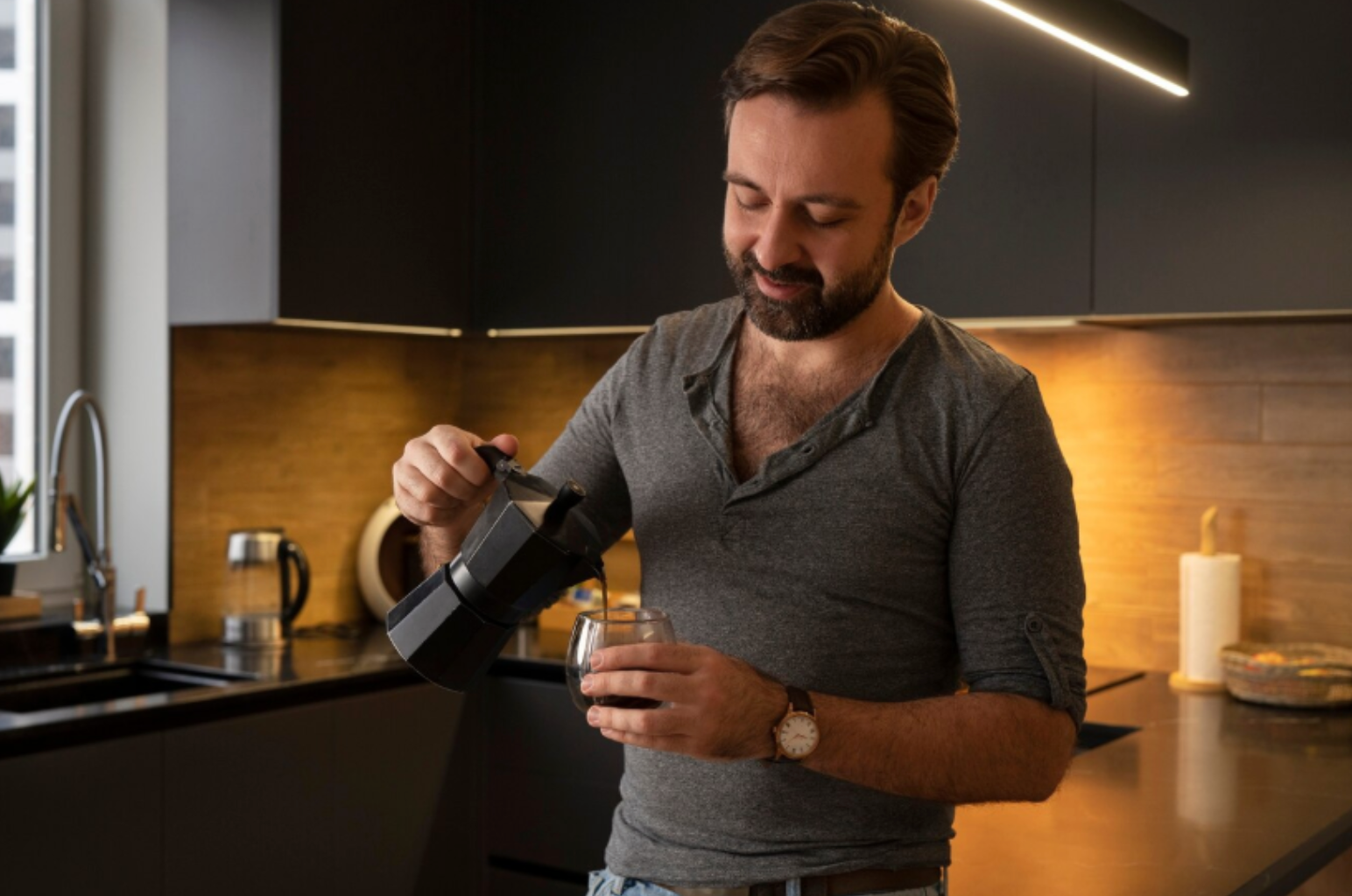 Man in a cozy kitchen pouring coffee from a moka pot into a glass, starting his morning routine