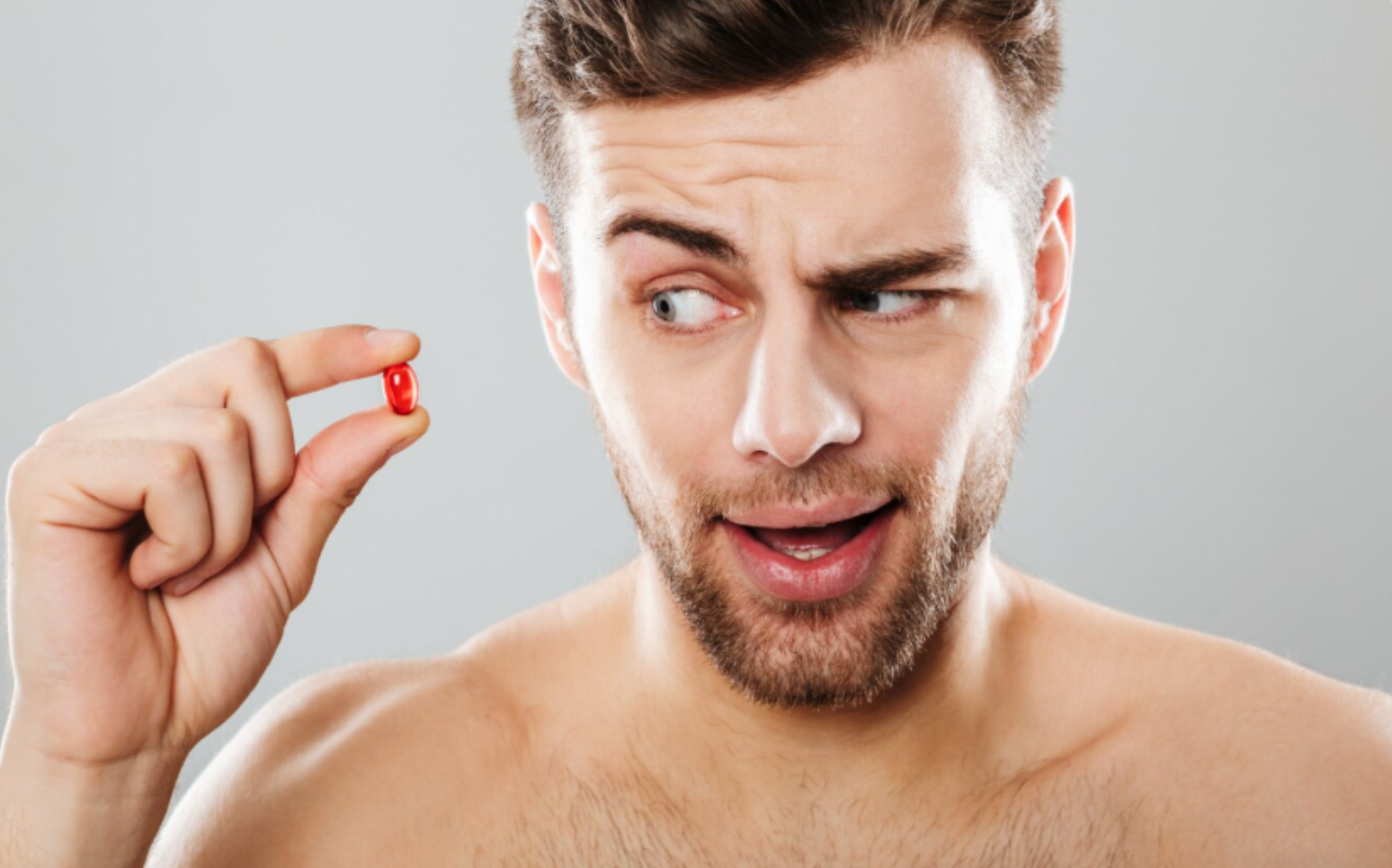 Man examining a red vitamin capsule with a skeptical expression, representing questions around hair supplement effectiveness.