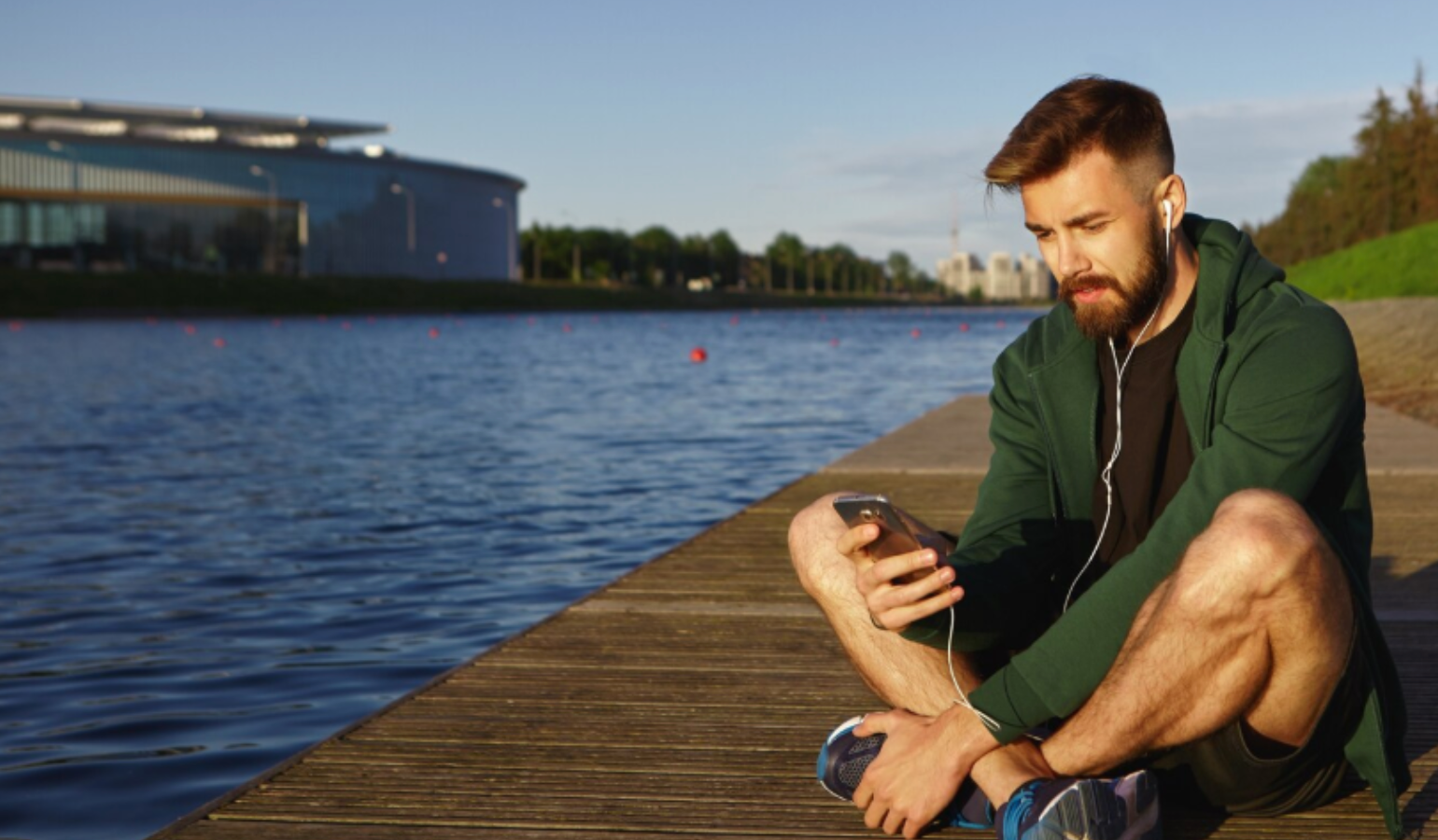Young man in athletic wear sitting by a lake, listening to headphones and looking at his phone during a calm morning workout. Represents focus, energy, and daily wellness habits.