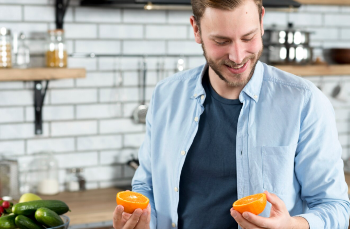 Man in light blue shirt smiling while holding two orange halves in a modern kitchen, representing natural food support for resveratrol supplement absorption.