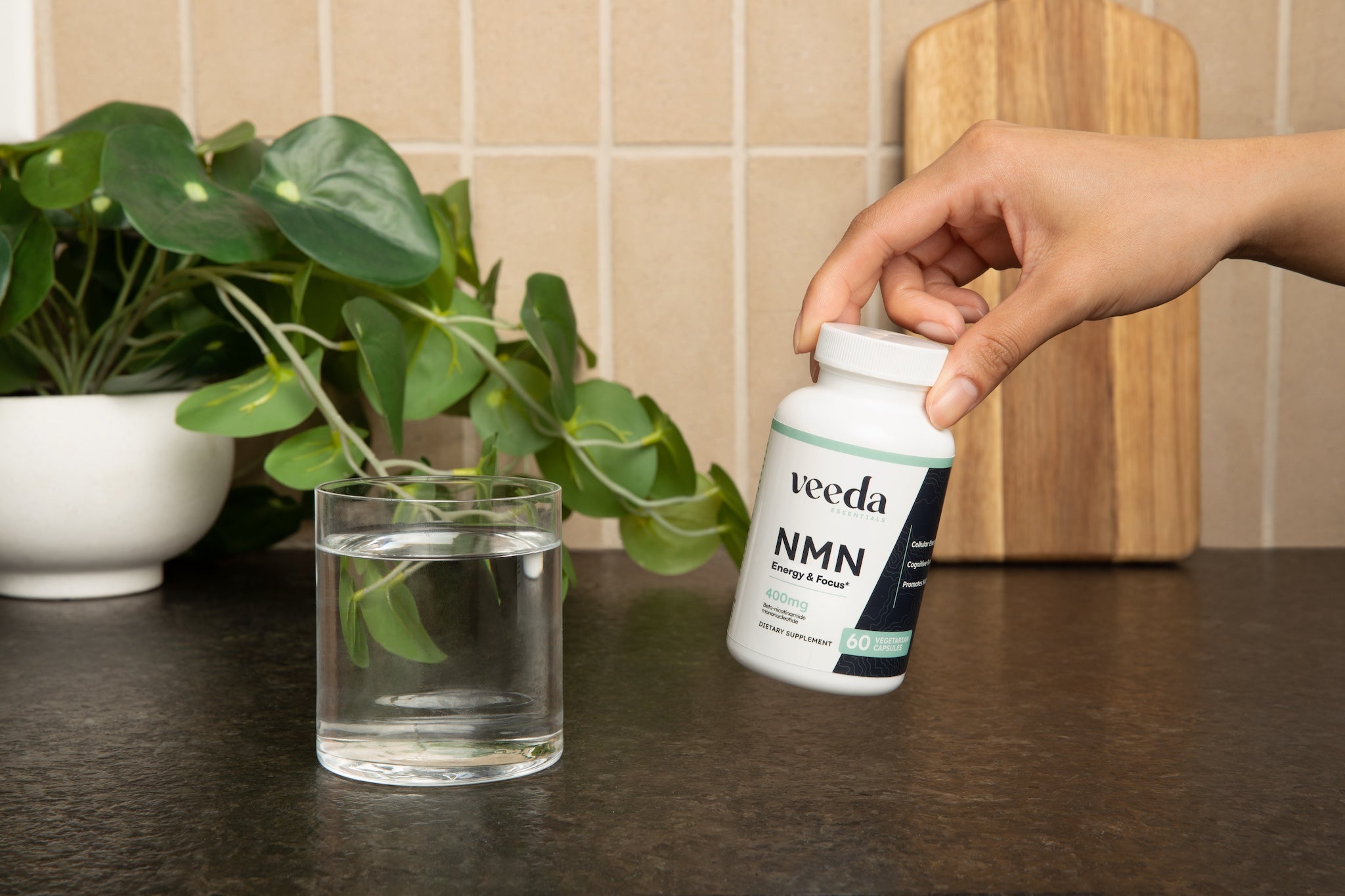 Hand holding a Veeda NMN supplement bottle on a kitchen counter with a plant and glass of water in the background.